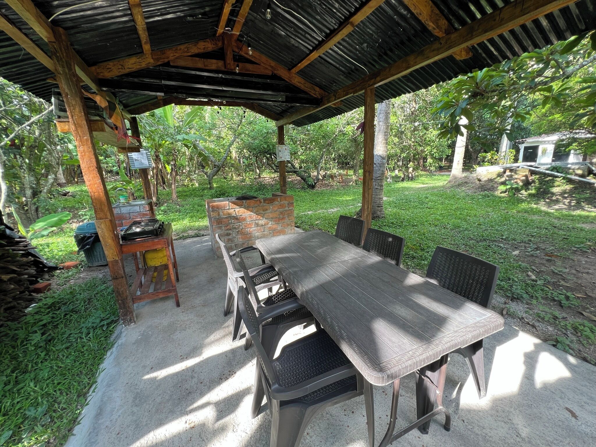 An outdoor covered patio with a table and chairs, surrounded by greenery and trees.