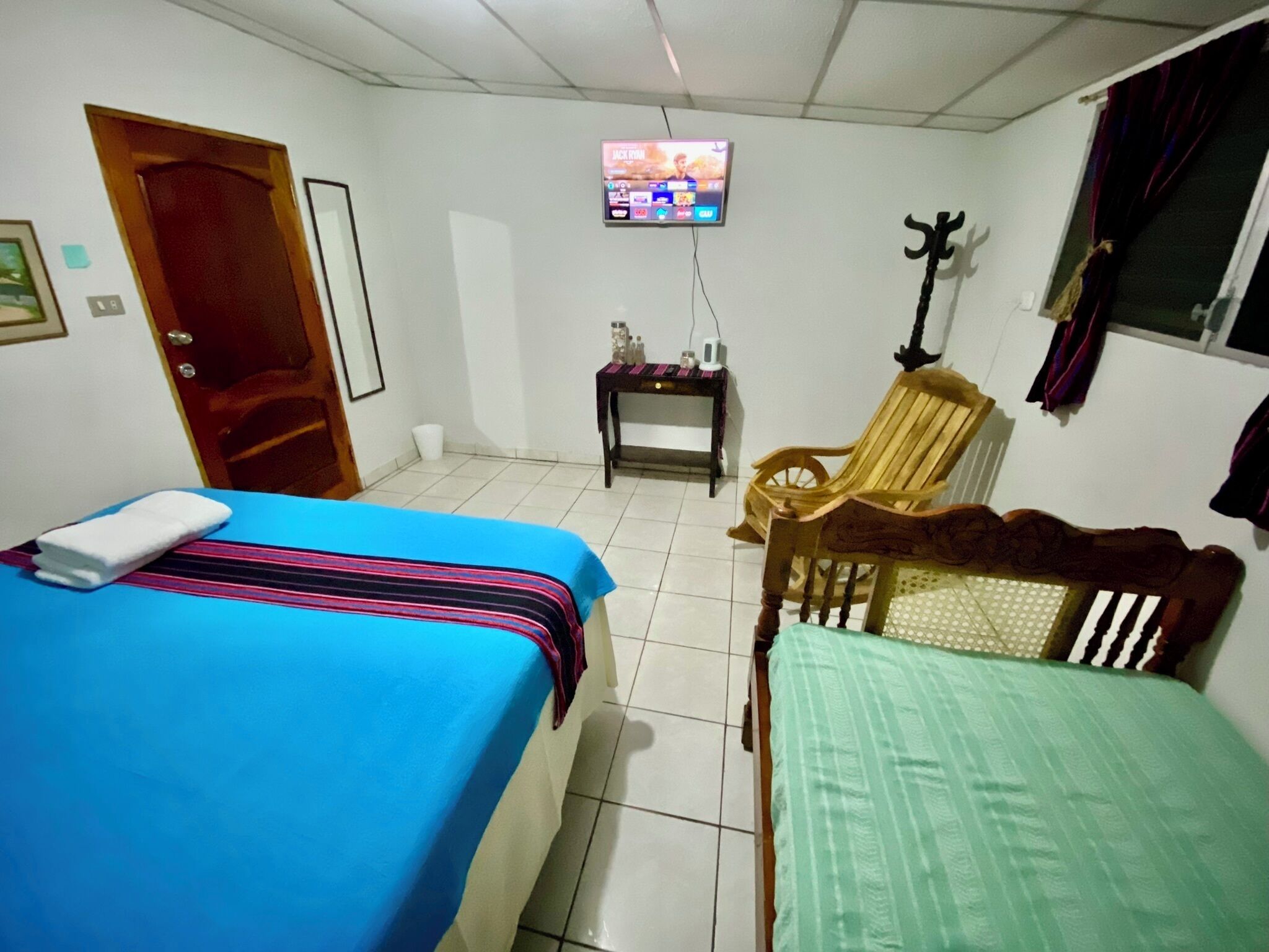 Quarto com duas camas, televisão presa na parede, uma cadeira de madeira e uma mesinha com abajur.