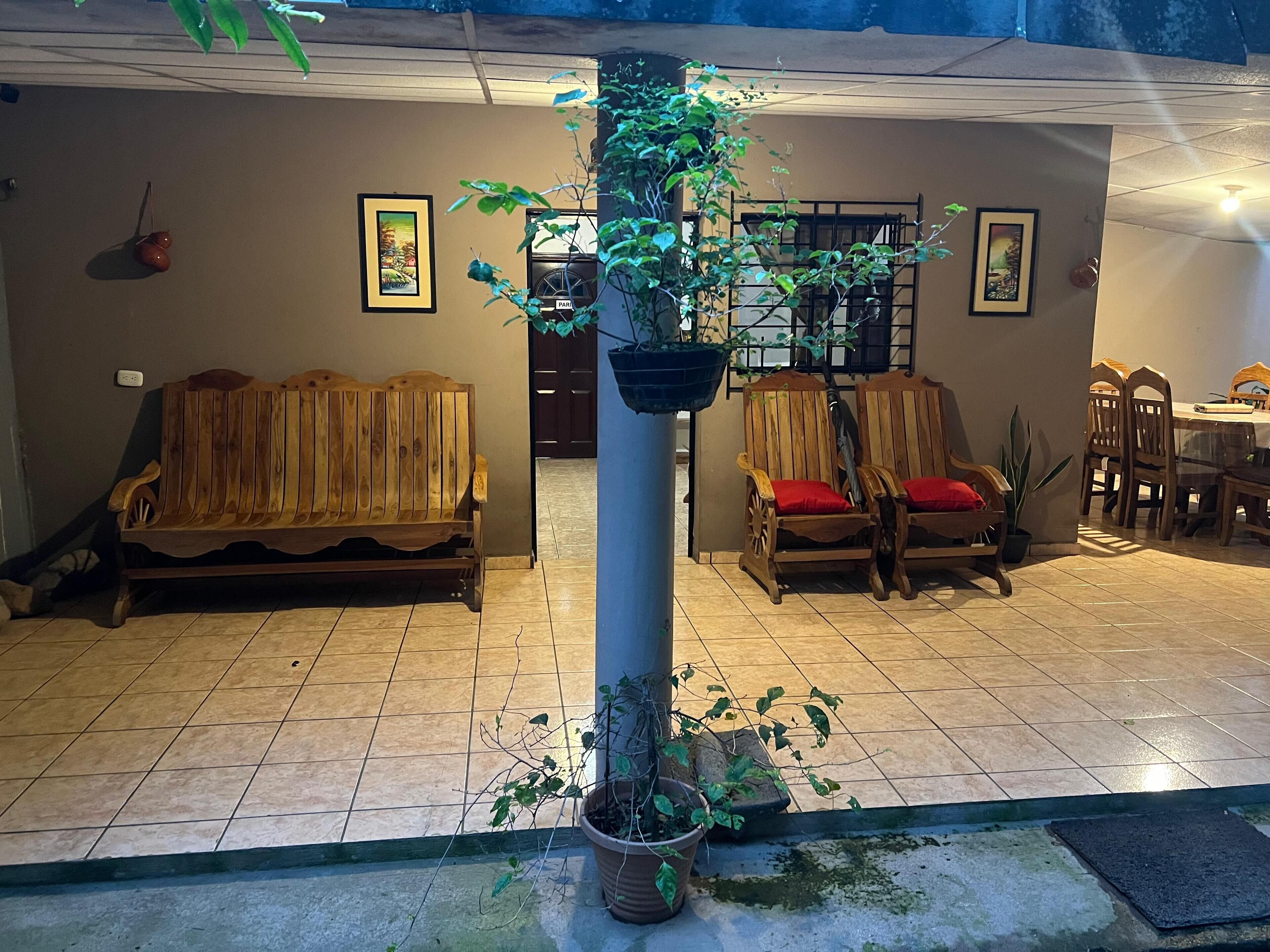 Un espace extérieur abrité, doté de bancs et de chaises en bois, de plantes en pot et d’un sol carrelé.