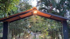 A wrought iron gate with decorative elements and lights, set in a lush garden with trees and plants. - Nanda Parbat Hostal (San Rafael Cedros)
