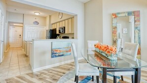 A modern kitchen with white cabinets, a refrigerator, and a microwave. There is a dining area with a glass table and chairs, and a painting on the wall. - Full Ocean View - SERENE SANDCASTLE 2 Bed/2 Bath (Gulfport)