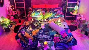 Una habitación con una cama cubierta por un edredón de temática tropical, rodeada de plantas en macetas y objetos decorativos.