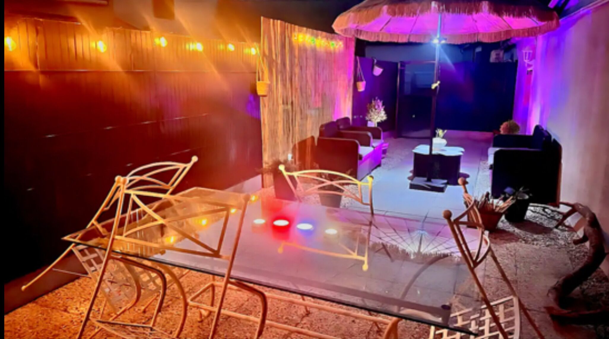 A room with a glass table, chairs, and a sofa. There are string lights and a curtain in the background.