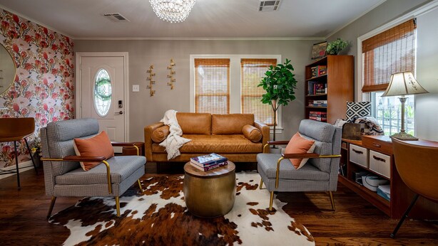 Un salon avec un canapé en cuir marron, des fauteuils à motifs, un tapis en peau de vache, un lustre et une bibliothèque.