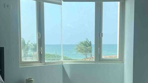 A bedroom with a bed, a nightstand, a lamp, and a large window with a view of the beach and ocean. - Trinity Beach Apt (Rio Grande)