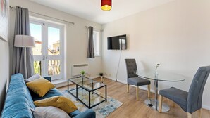Un salon moderne comprenant un canapé, une table basse en verre, une table à manger avec des chaises, un téléviseur fixé au mur et une fenêtre avec des rideaux.