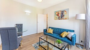 Un salon moderne avec une table basse en verre, un canapé bleu agrémenté de coussins jaunes, un fauteuil gris et un tableau représentant un éléphant.