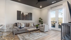 A modern living room with a sofa, coffee table, and chairs. There is a balcony with a view of trees. - 3 Bedroom, Main Level - ADA  (Helena)