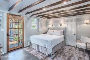 A bedroom with a large bed, wooden beams on the ceiling, a door with a glass panel, and two wall-mounted lamps. - Relaxing garden cottage with sauna
(Shreveport)