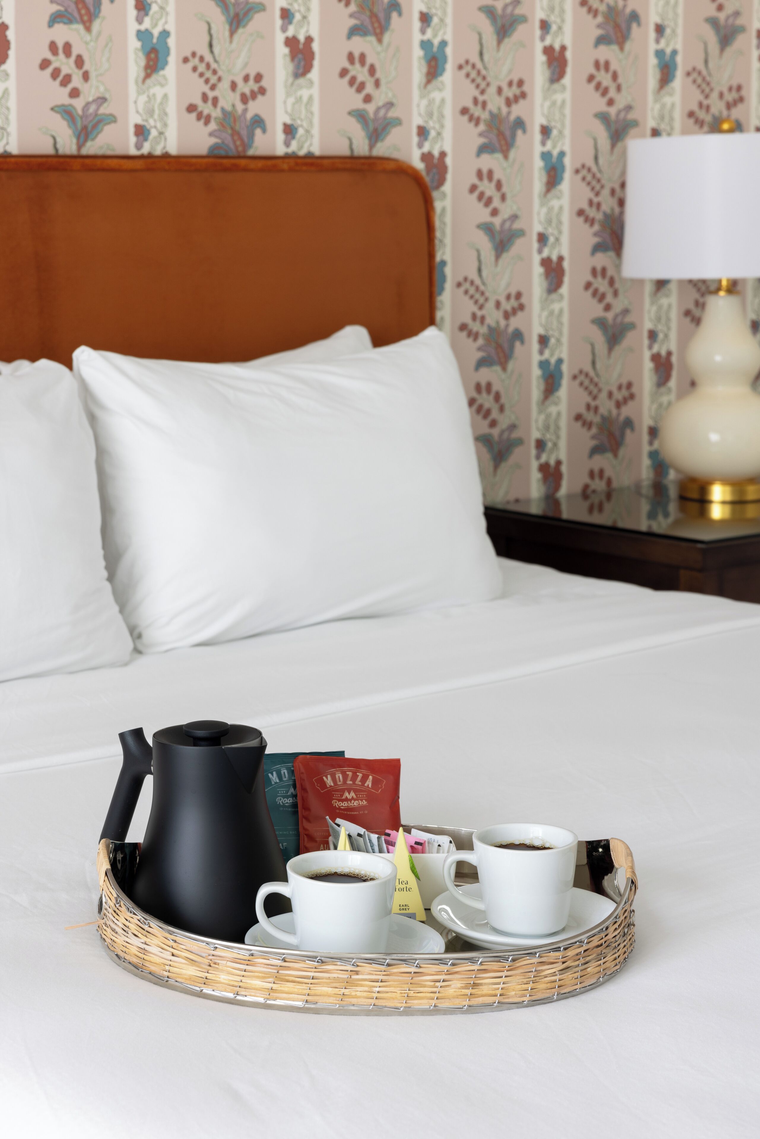 Una cama bien hecha con sábanas blancas, una tetera negra, dos tazas de café y una bandeja con una bolsita de café y una bolsita de té.