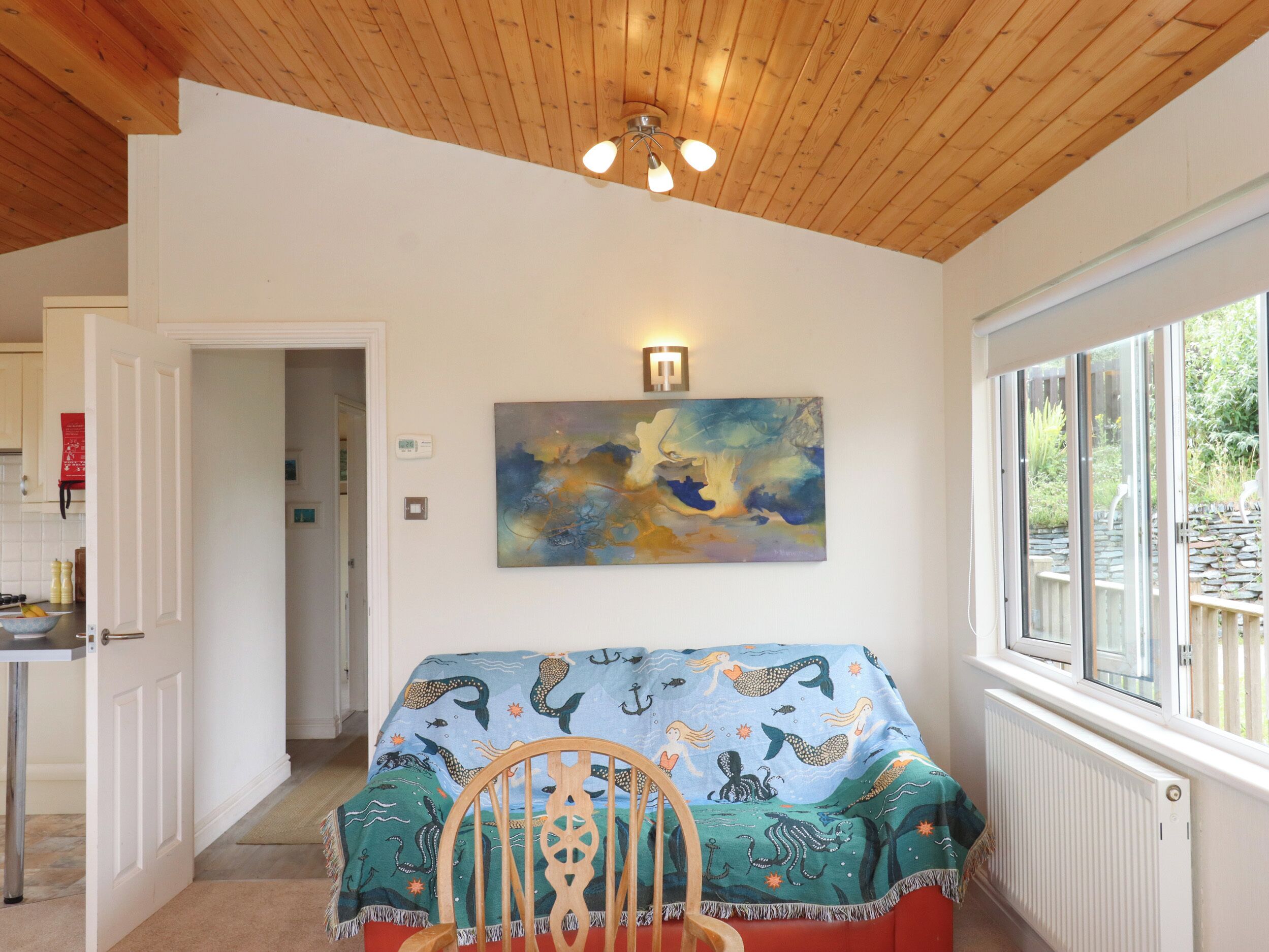 A cozy room with a wooden ceiling, a colorful abstract painting, a chair with a mermaid-themed blanket, and a view of a garden through a window.
