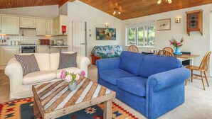 Una sala de estar acogedora con un sofá azul, un sofá blanco y una mesa de centro de madera con un jarrón de flores. Se aprecia una cocina al fondo con armarios blancos y una estufa.