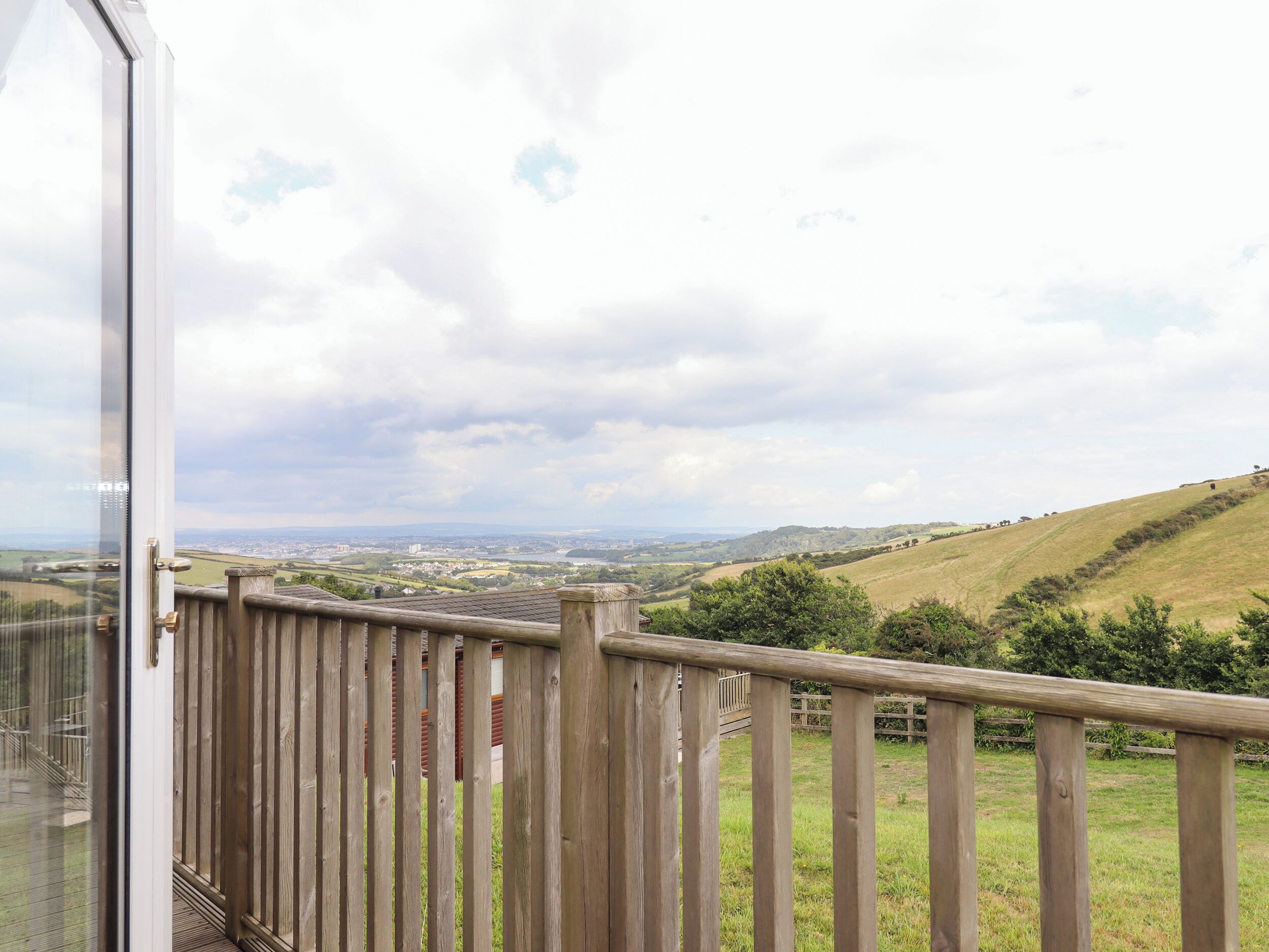 A wooden balcony with a glass door, overlooking a rural landscape with hills and a distant town.