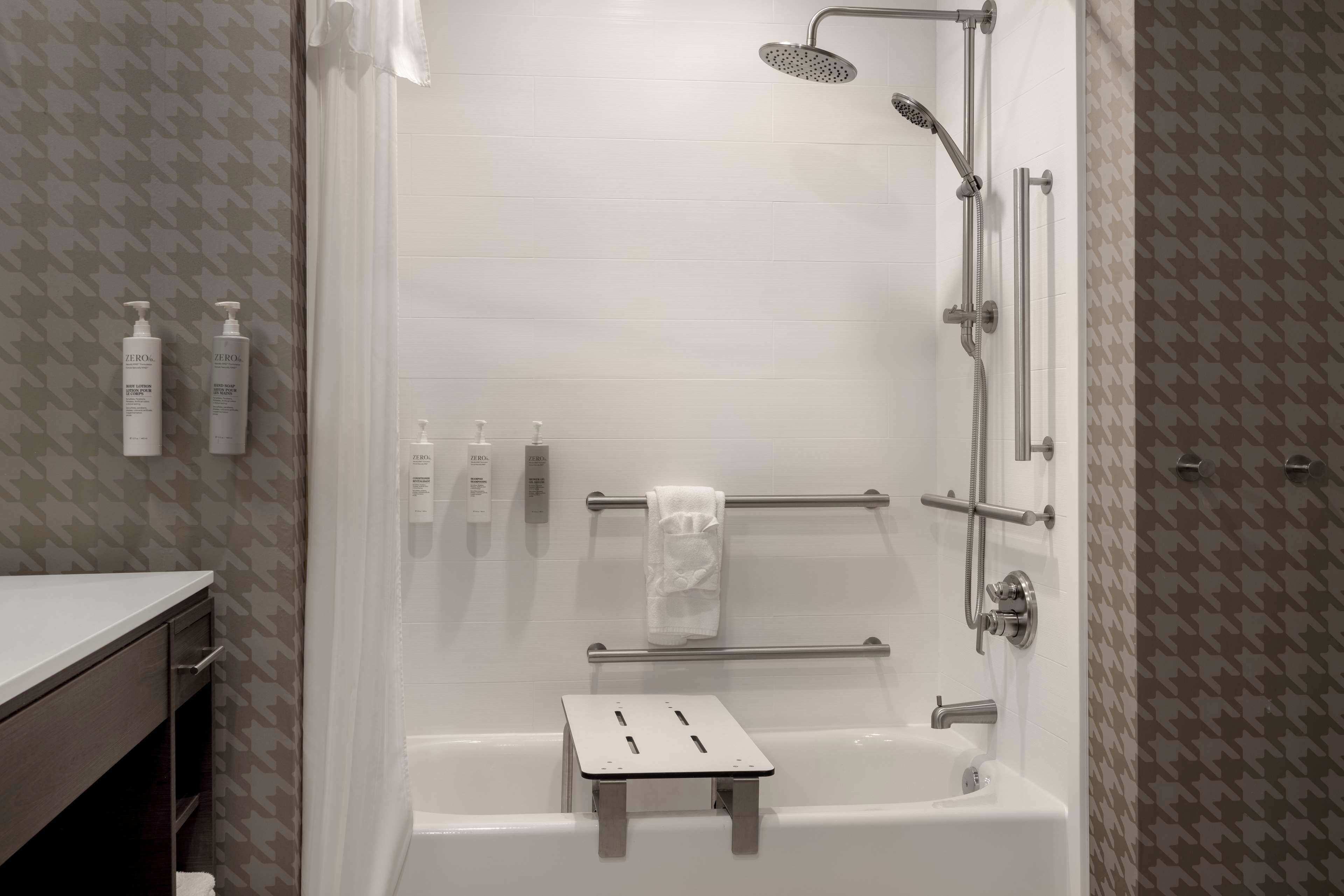 A modern bathroom with a bathtub, shower, and handrails. There are two bottles of hand soap on the wall.