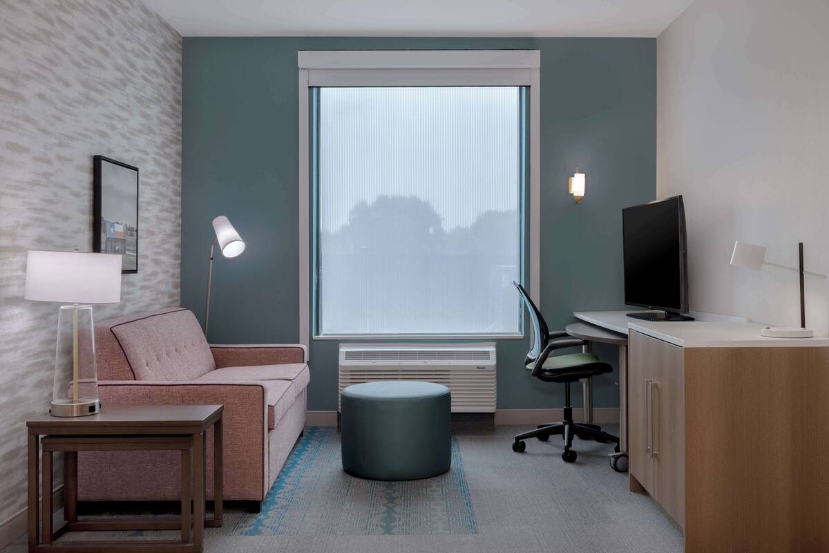 Een moderne hotelkamer met een bank, een bureau met een computer, een televisie en een groot raam met zonwering.