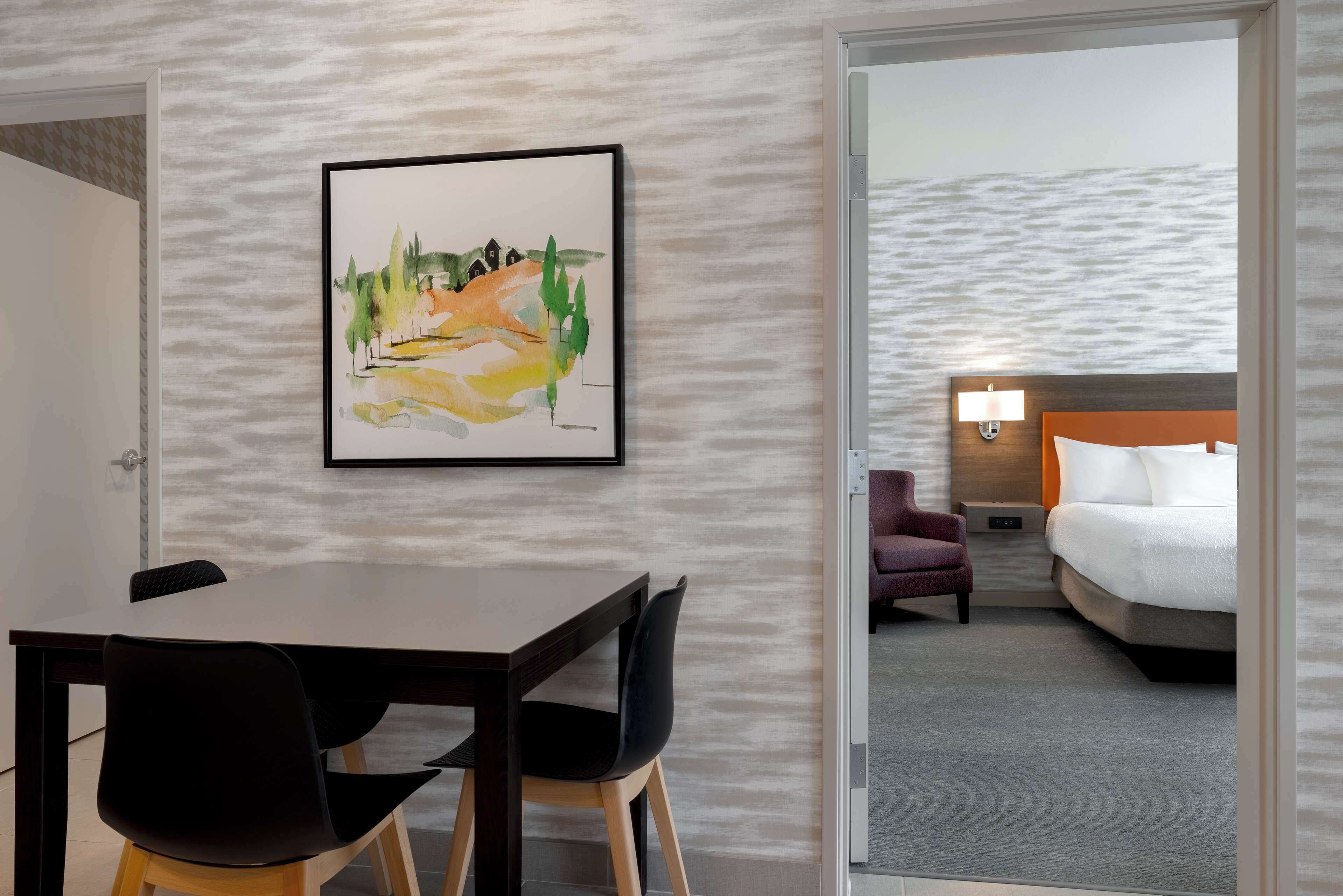 A hotel room with a bed, a nightstand, a lamp, a chair, and a painting on the wall.