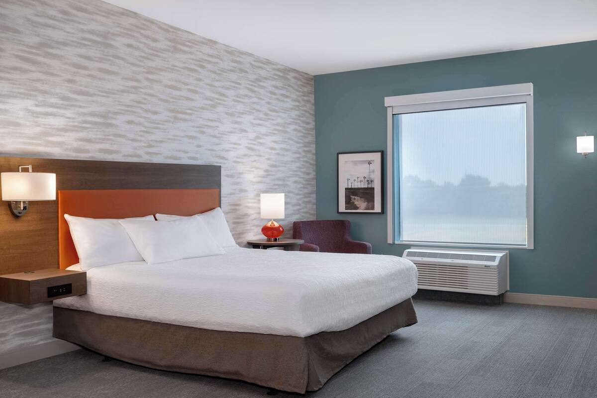 Een hotelkamer met een groot bed, een nachtkastje met een lamp, een stoel, een raam met jaloezieën en een luchtverfrisser aan de muur.