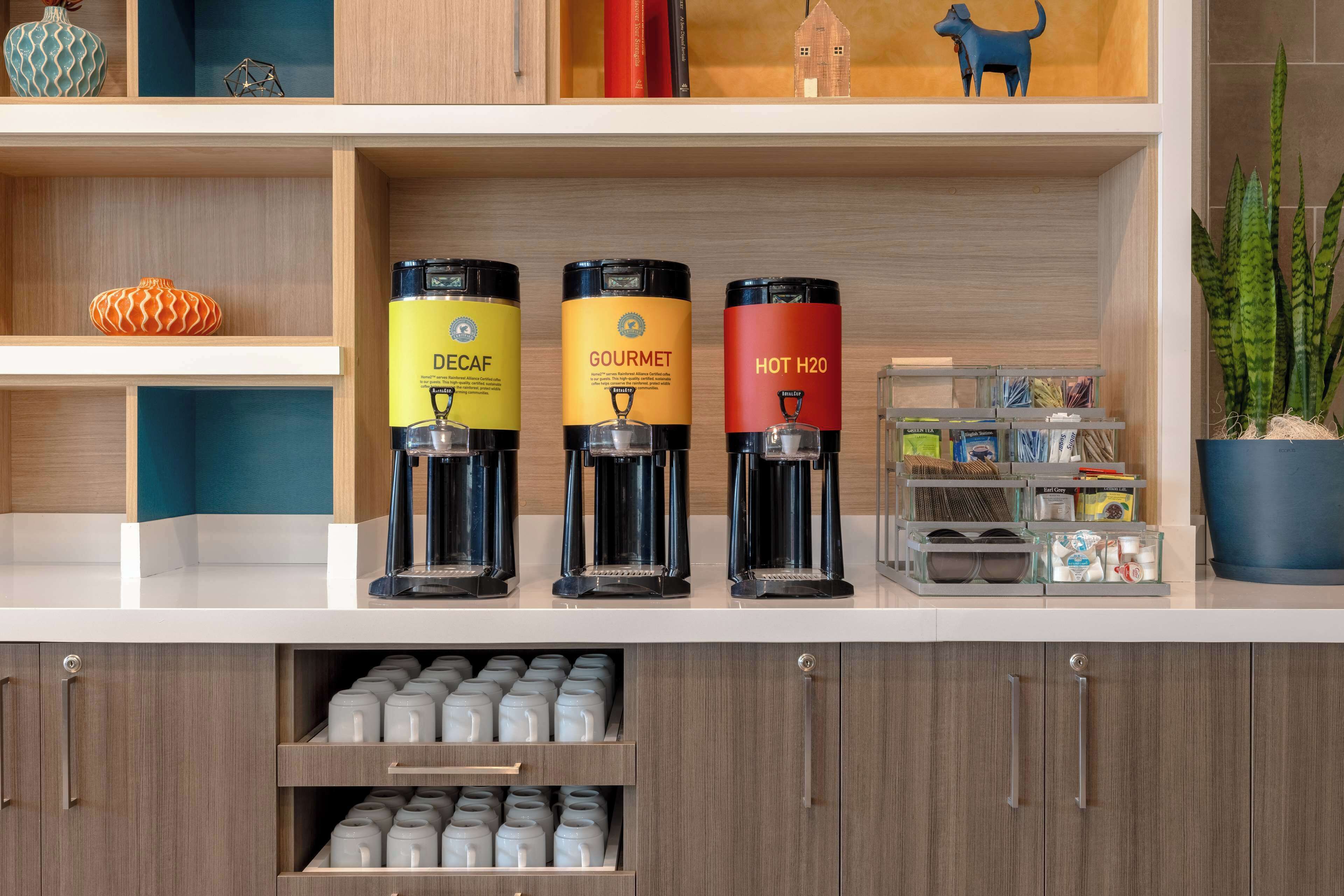 Eine Küchenarbeitsplatte mit drei Kaffeemaschinen, beschriftet mit 'Decaf', 'Gourmet' und 'Heißes Wasser'. Darunter befinden sich Schränke mit Vorratsgläsern.
