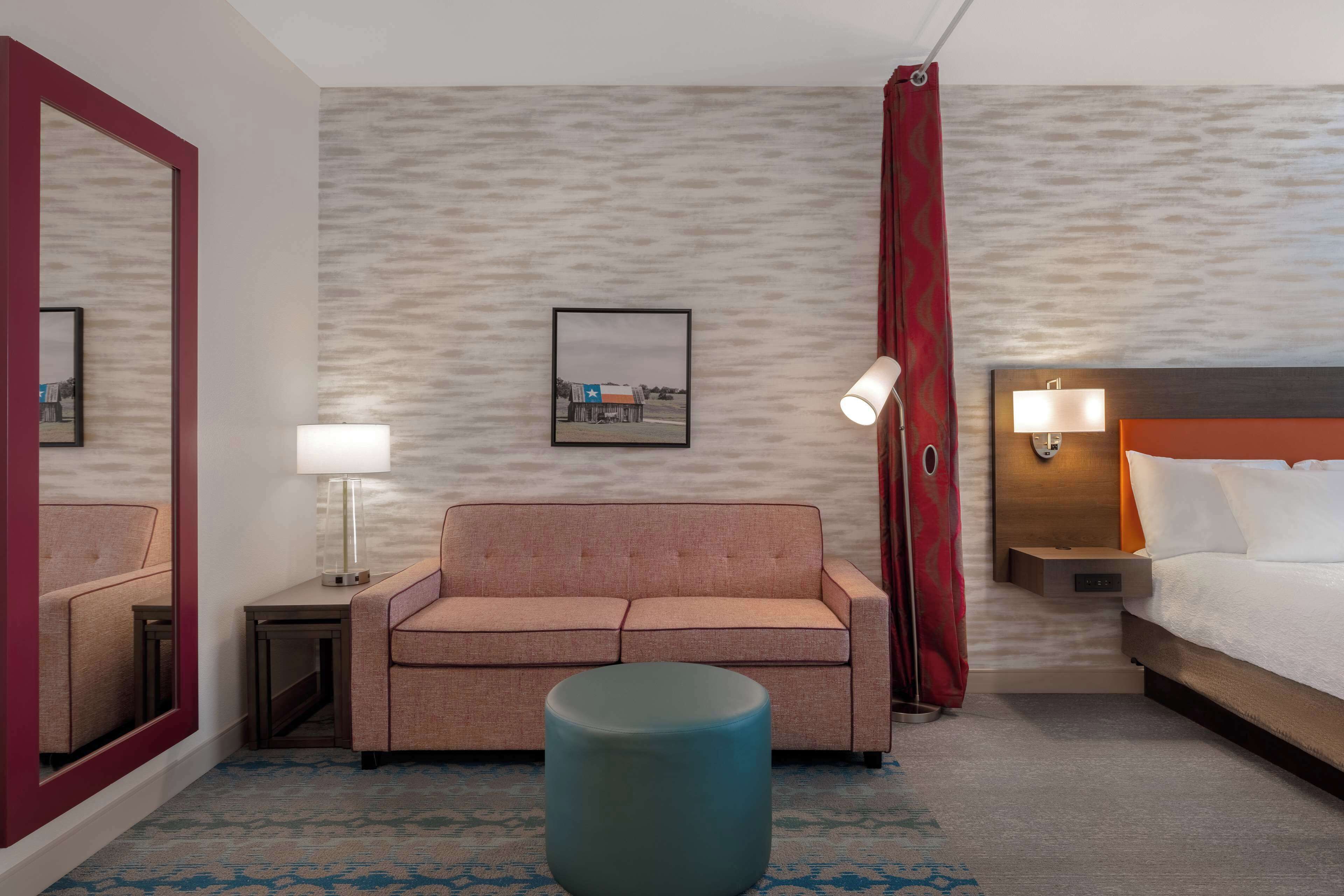 Una habitación de hotel con sofá, una mesa redonda, cama, mesita de noche con lámpara, un cuadro en la pared y un espejo.