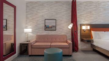 Una habitación de hotel con sofá, una mesa redonda, cama, mesita de noche con lámpara, un cuadro en la pared y un espejo.