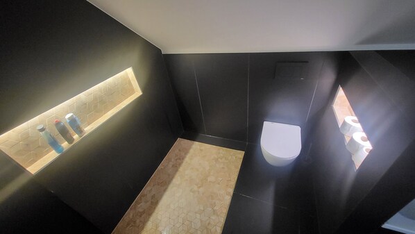 Un baño moderno con paredes oscuras, suelo de madera, un inodoro y un estante con botellas.
