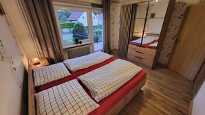 A bedroom with a bed, a dresser, a mirror, and a window with a view of a house and greenery.