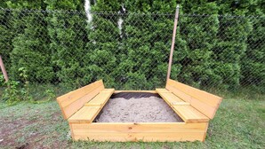 A wooden raised garden bed with a black bottom, surrounded by a chain-link fence and dense green foliage in the background.