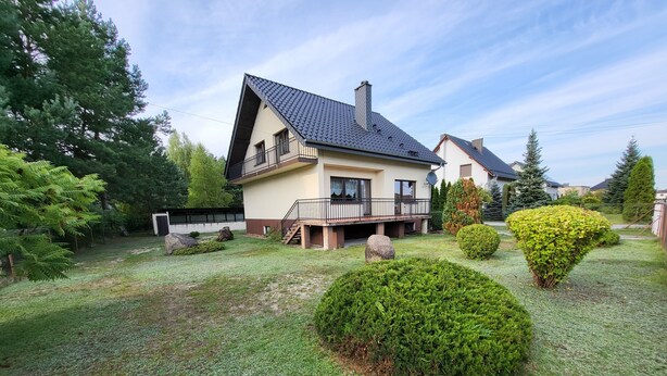 A modern two-story house with a sloped roof, a balcony, and a well-maintained lawn with bushes and a rock feature.