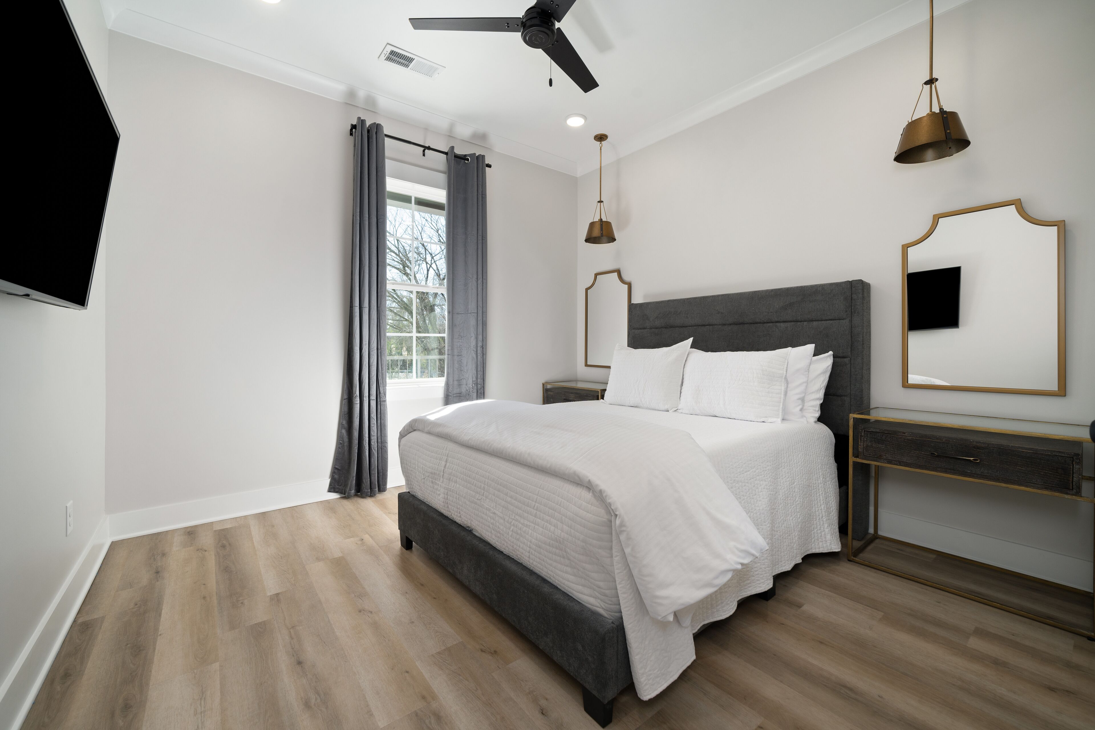 A modern bedroom with a large bed, a ceiling fan, a bedside table, a nightstand, a TV, and a window with curtains.