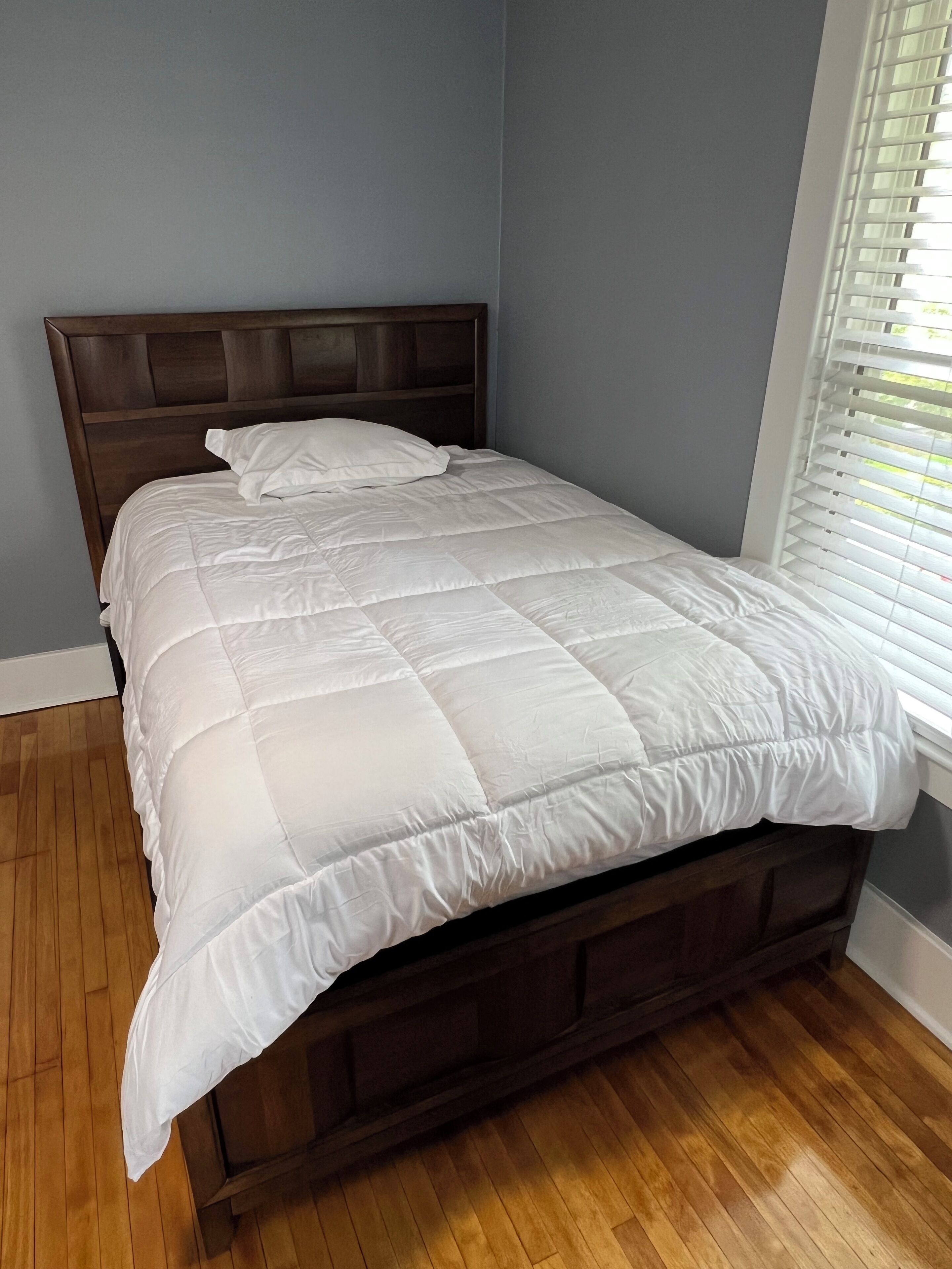 A bed with a wooden headboard and a white quilt in a room with wooden flooring and a window with blinds.