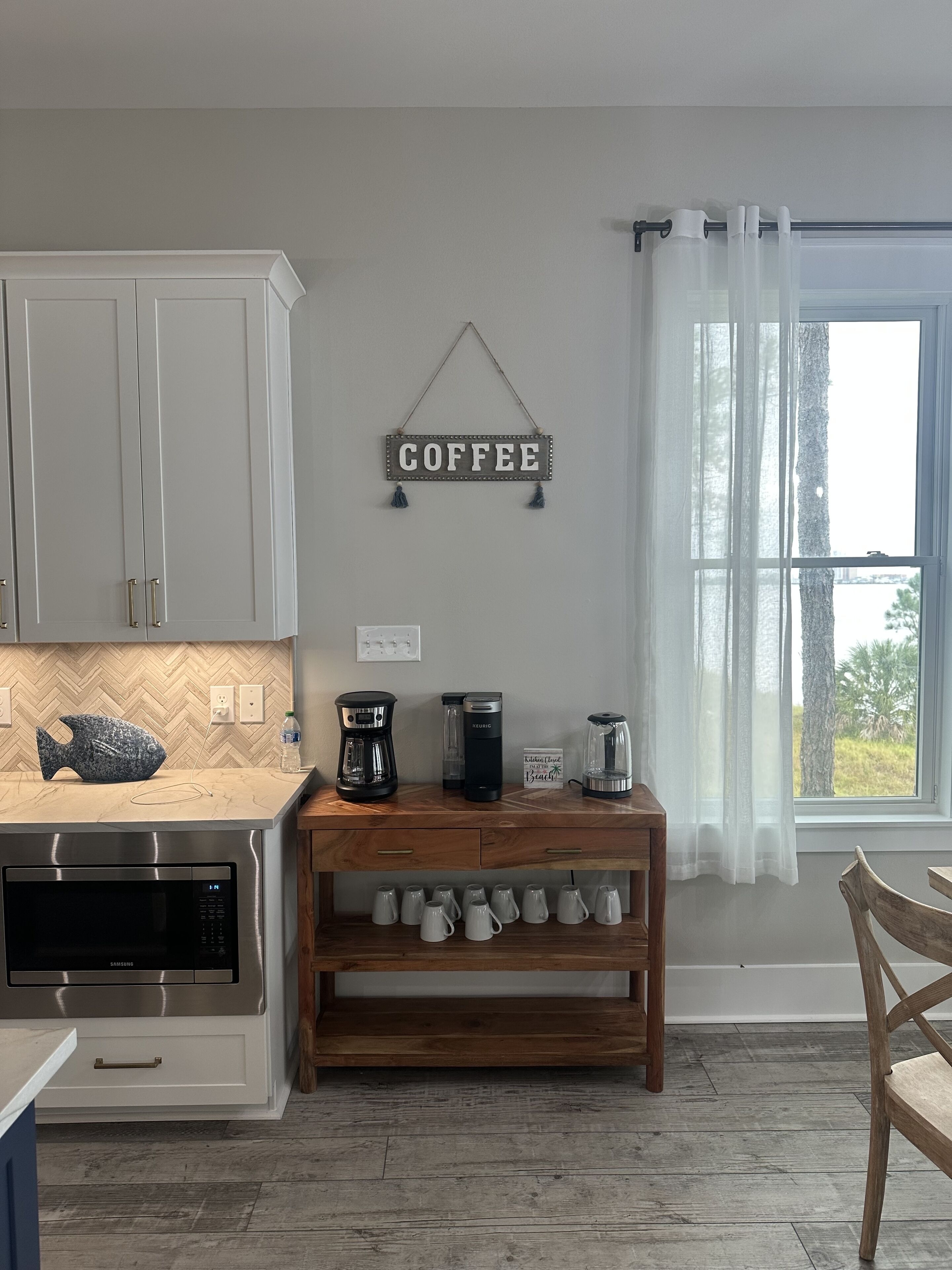 Cozinha com armários brancos, um letreiro de café, uma cafeteira, um liquidificador e uma estante para canecas de café.