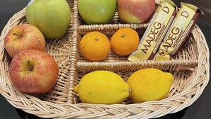 A wicker basket containing apples, oranges, lemons, and Madeg Good brand bars.