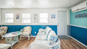 A room with a blue and white color scheme, featuring a white wicker sofa, a small round table, and wicker chairs. There are framed pictures on the wall and a sign that reads "BEACH COTTAGE".