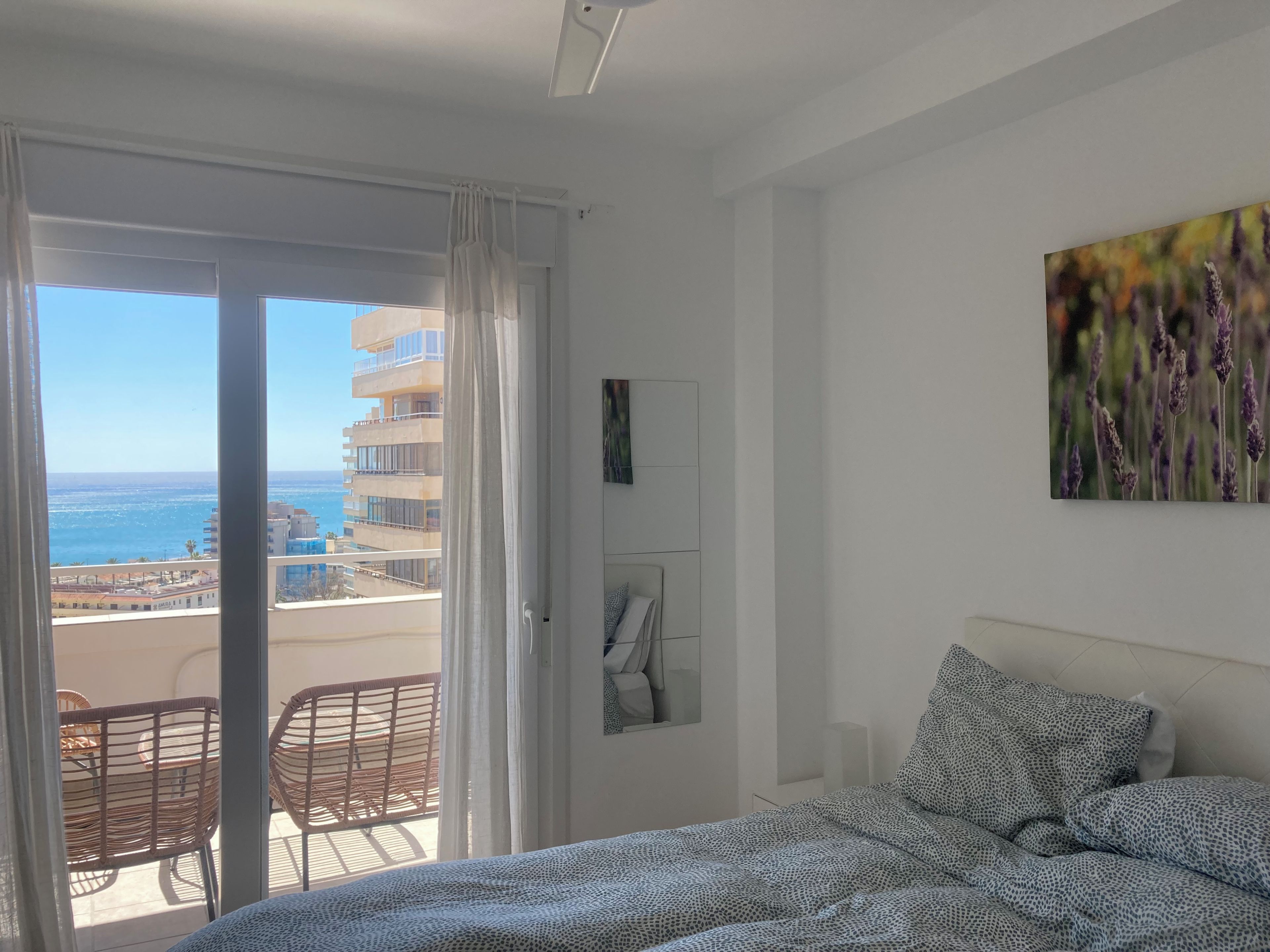Un dormitorio con cama, un balcón con sillas de mimbre, un cuadro y vista al mar y edificios.