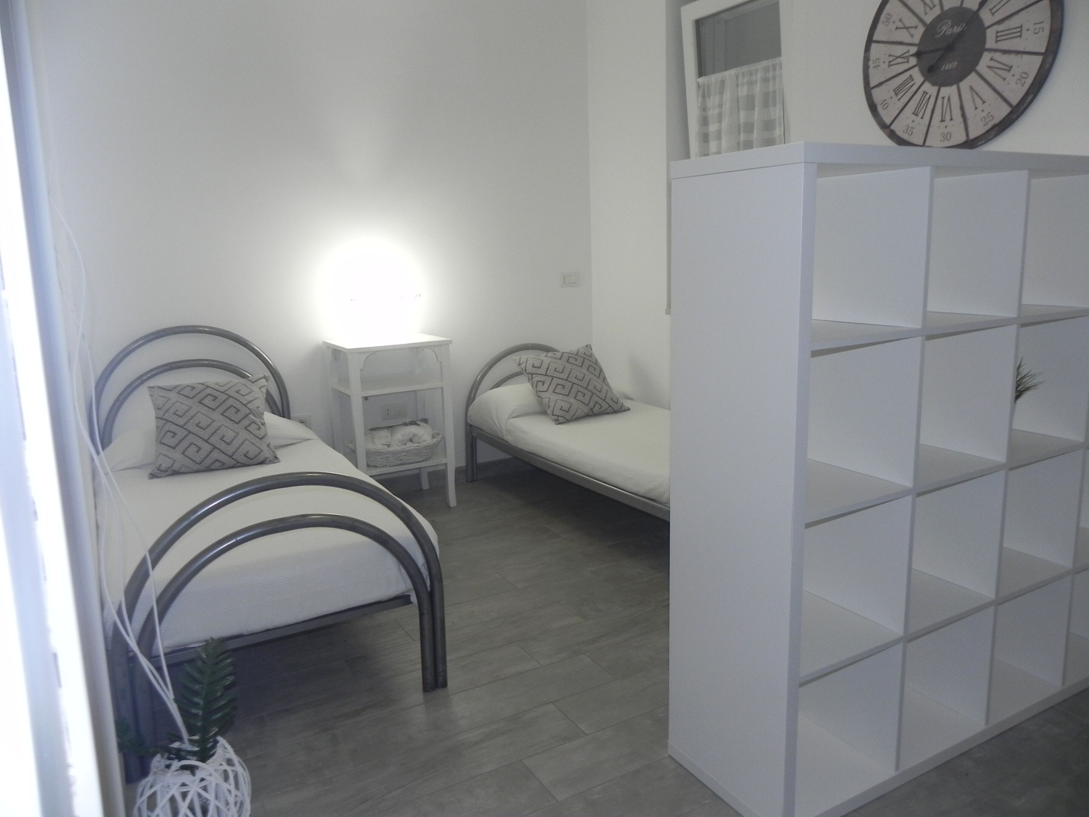 Quarto com duas camas, uma pequena mesa, estante e relógio de parede.