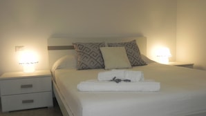 Una cama bien tendida con una colcha blanca, dos almohadas estampadas y una toalla doblada con una flor de lavanda. Hay dos lámparas de noche sobre mesitas blancas.
