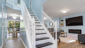 Uma escadaria leva a um segundo andar, com área de refeições, cadeiras e mesa, sala de estar com sofá e televisão de tela plana, e uma lareira.