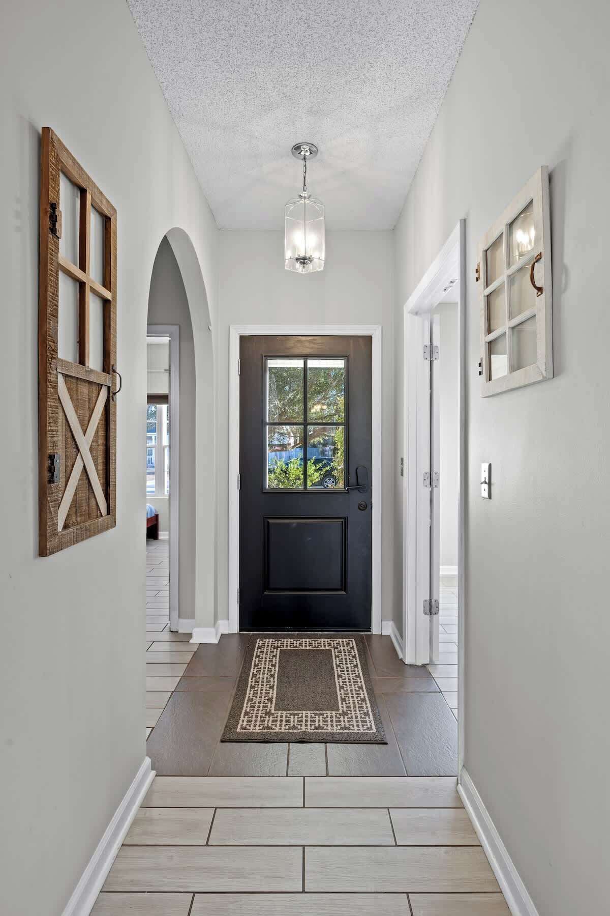 A hallway with a dark front door, a light-colored ceiling, and a patterned rug leading to a room.