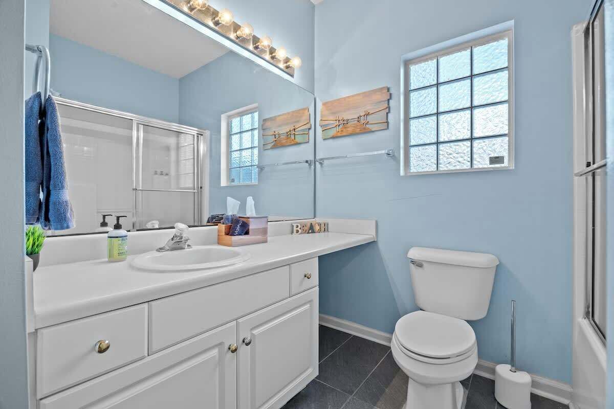A bathroom with a white vanity, a sink, a mirror, and a toilet. There are two windows with frosted glass panes.