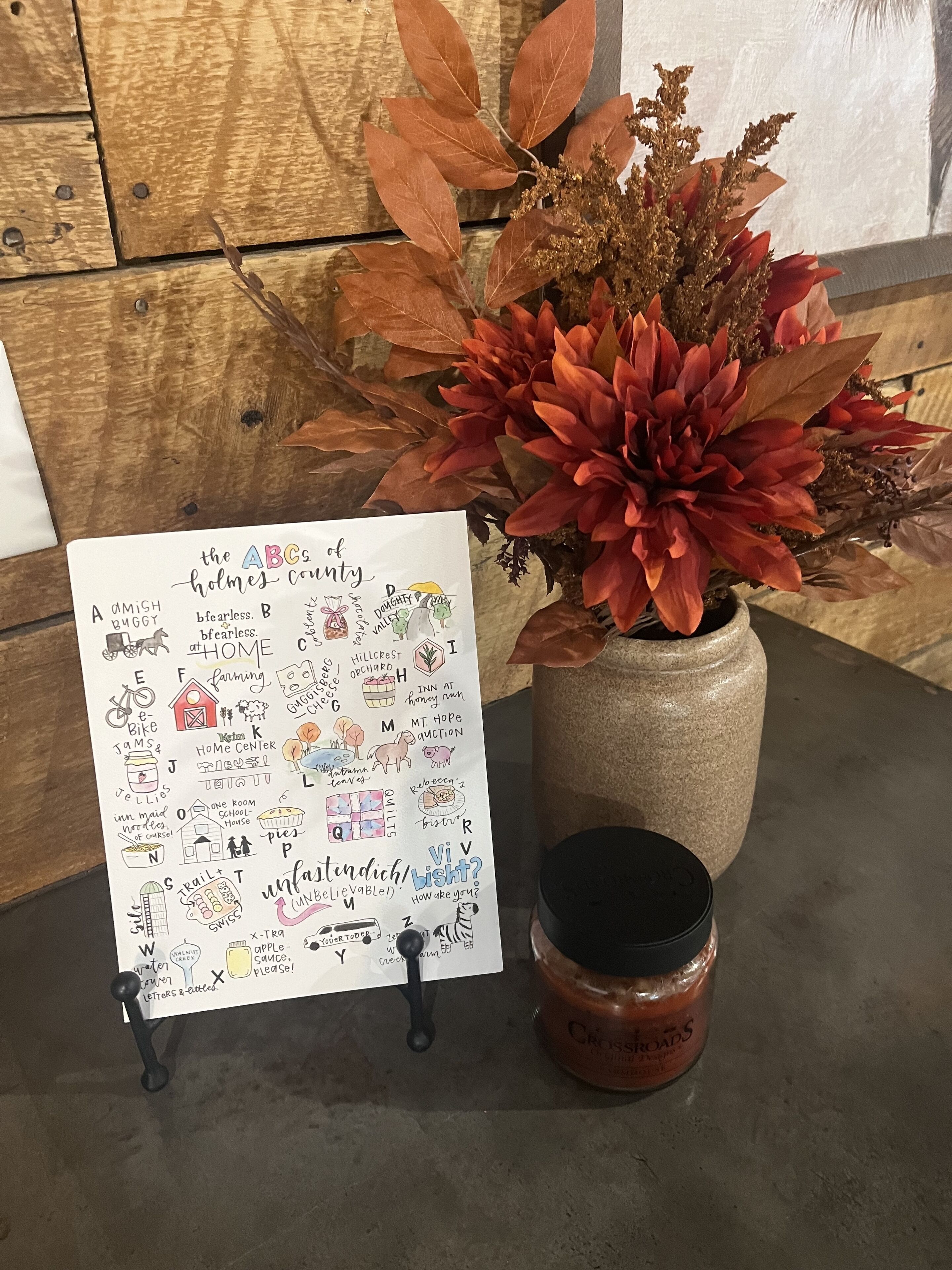Un cartello scritto a mano con scarabocchi e scritte varie, un vaso con fiori rossi e un vasetto di senape e miele.