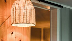 Une porte en bois ornée d’un luminaire tressé, un lit visible avec une tête de lit blanche, et une porte coulissante entrouverte révélant une pièce avec un lit et une table de chevet.