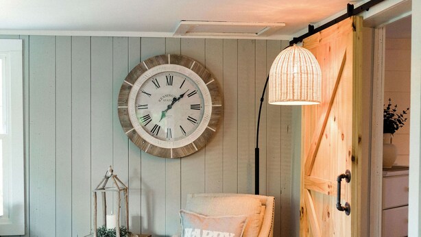 A cozy room with a wooden floor, a white armchair with a 'Happy' pillow, a wall clock, a lantern, and a wooden door.