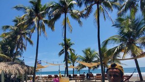 A poolside area with palm trees, a thatched-roof structure, and a person holding a tray with drinks. - Beachfront Casa Maya resort (Tamanique)