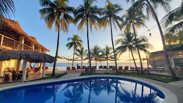 A poolside area with palm trees, lounge chairs, and a thatched-roof structure.