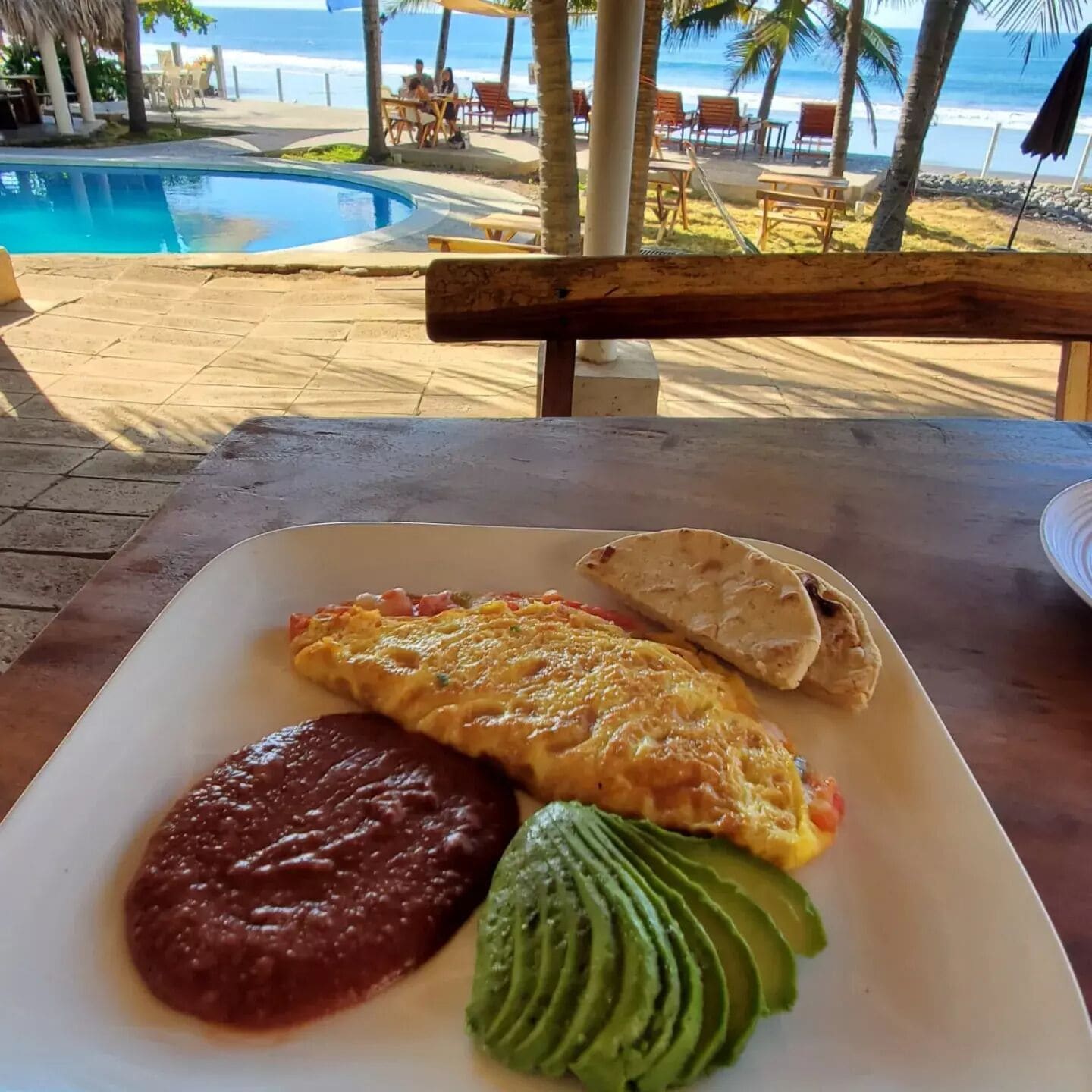 A dining table with a plate of food, a pool, and beach chairs in the background.