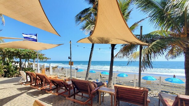 A beachside seating area with wooden loungers and blue umbrellas, overlooking the ocean. - Beachfront Casa Maya resort (Tamanique)