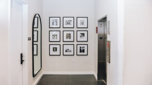 A hallway with a series of framed photographs on the wall, a large mirror, and an elevator. - The Neighborhood Hotel - Little Italy Two Bedroom (Chicago)