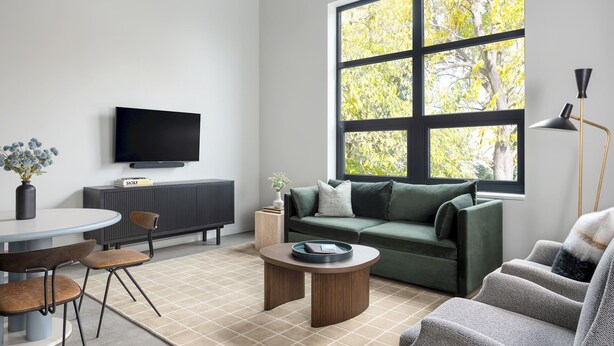 Een moderne woonkamer met een groot raam, een flatscreen televisie, een groene bank, een salontafel, en een eethoek met stoelen en een tafel.