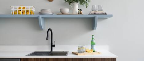 A kitchen with a white countertop, a black faucet, a wooden cabinet, a white shelf with kitchen items, and a potted plant.