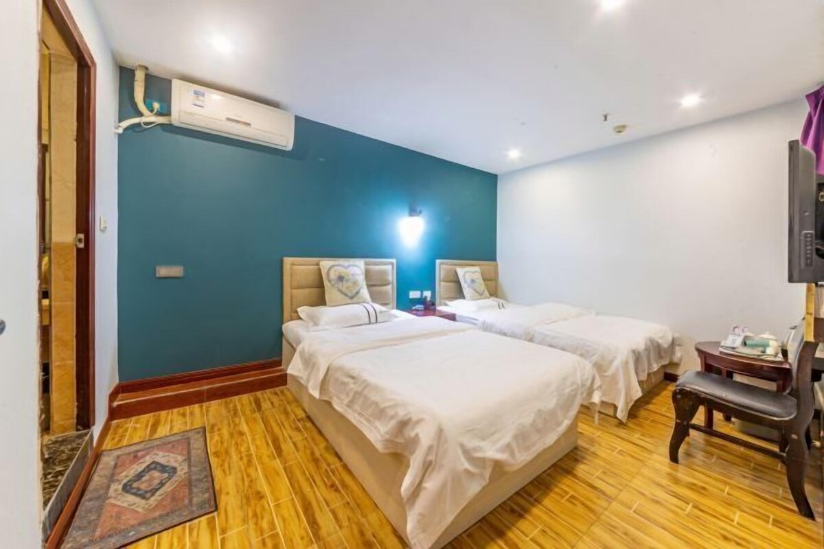 A hotel room with two beds, a wooden floor, a wall-mounted air conditioner, and a small table with a chair.